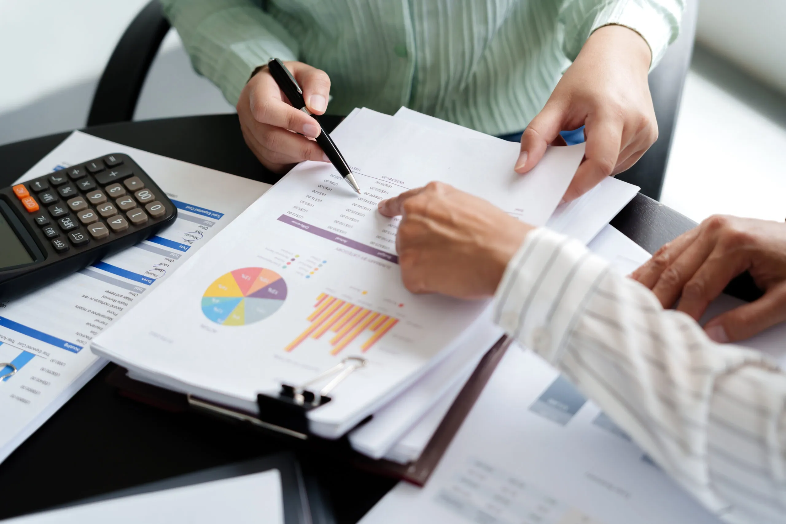Two people’s hands point at a financial report document on a black desk.