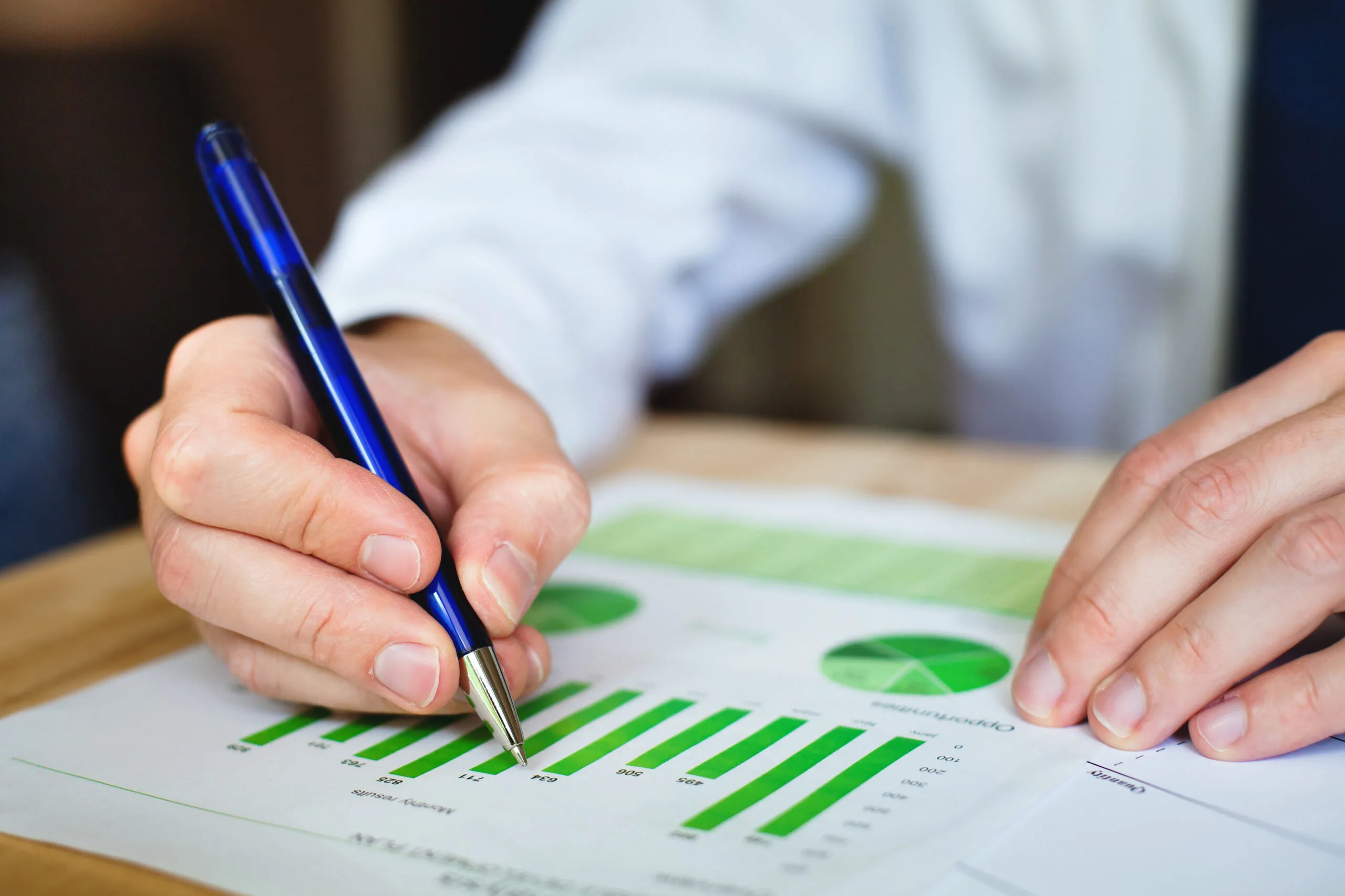 A person holds a blue ball-pointed pen while writing on a piece of paper with green graphs and charts on it.