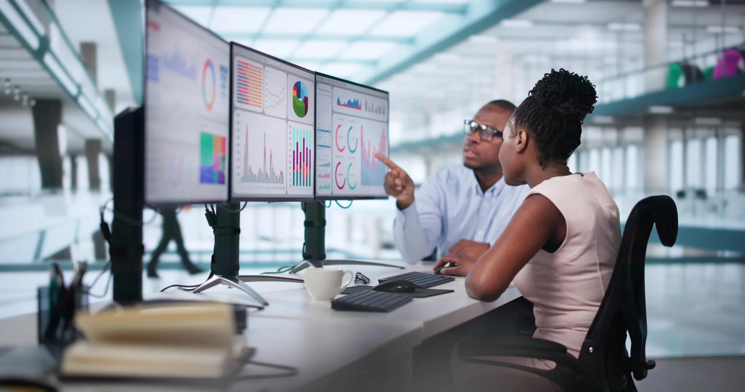 A man at a desk points at a computer screen with charts and graphs while a woman looks at the screen.
