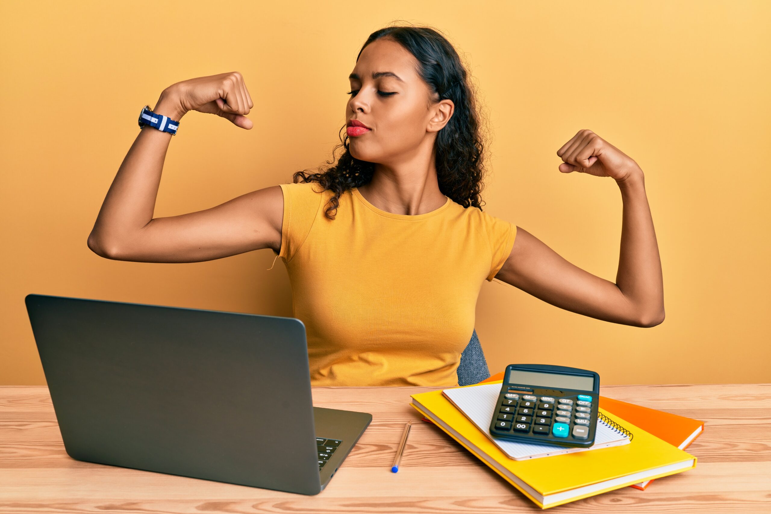 A woman in a yellow t-shirt flexes while looking at her right bicep.