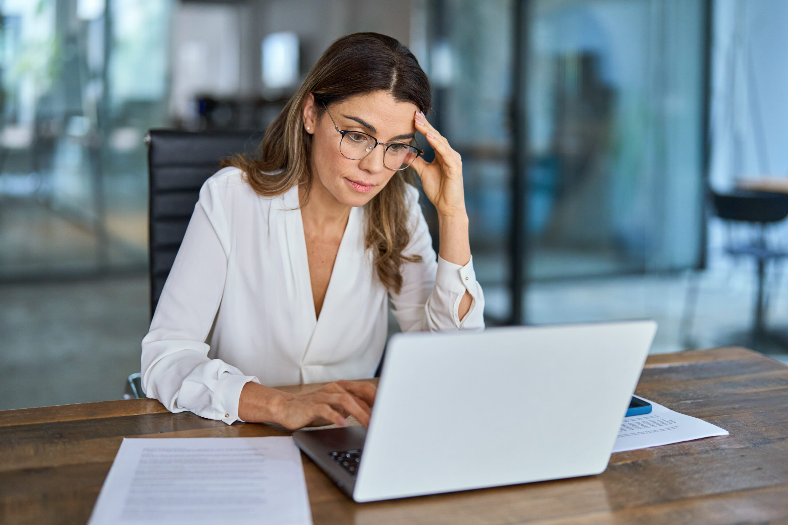 A woman in glasses places her hand on her head while looking down at her laptop on a wood desk.