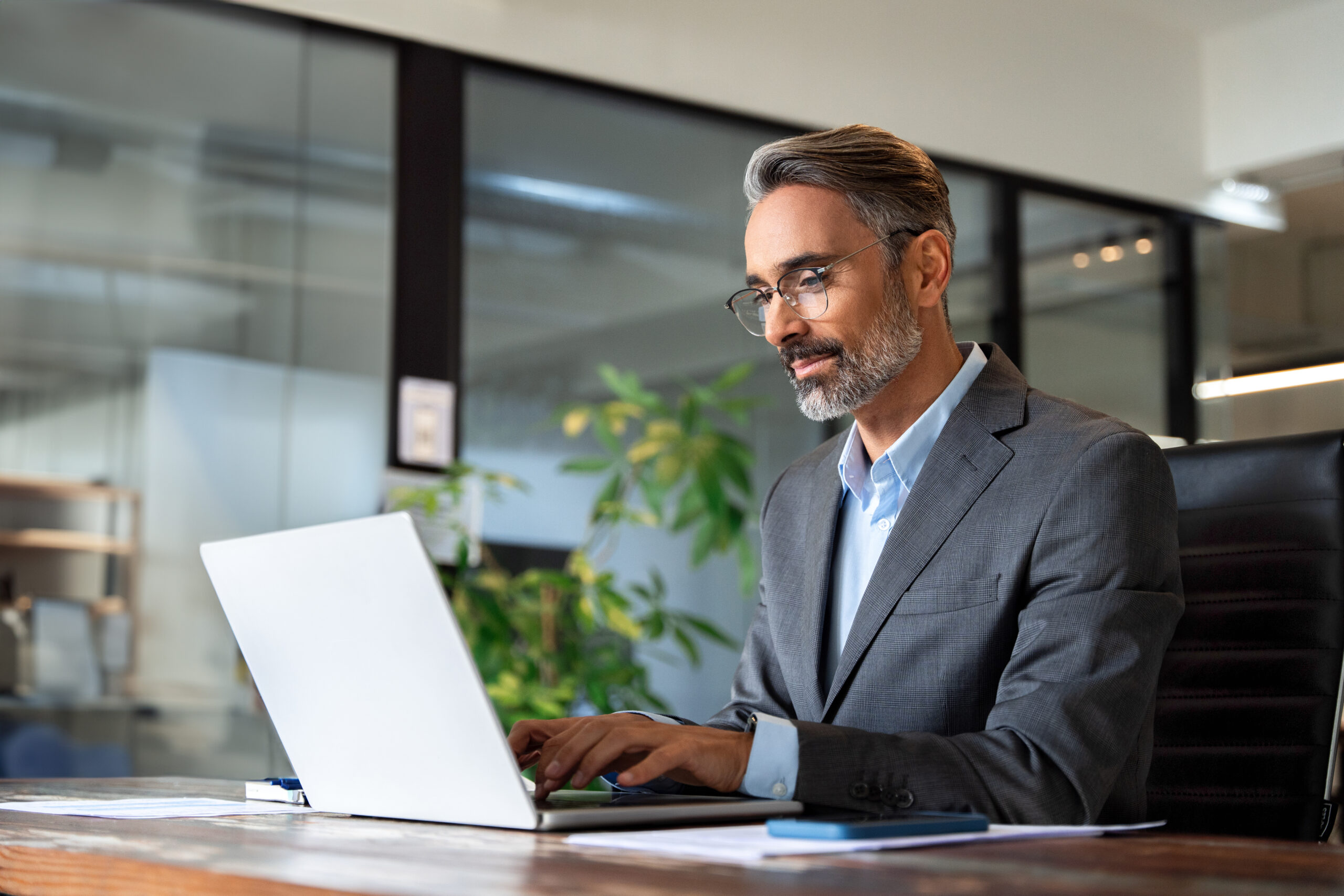 A man wearing a suit sits at a desk smiling while typing on his computer.