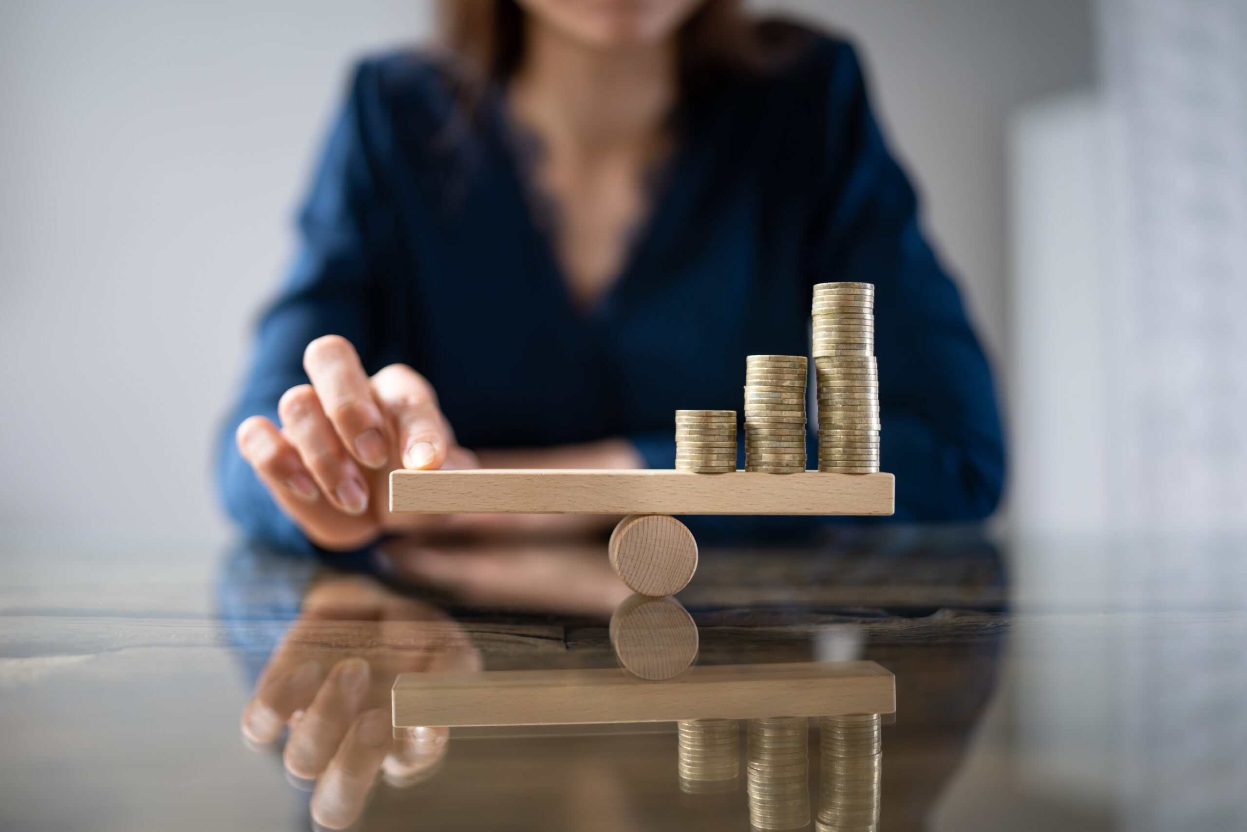 A woman presses on a wooden scale that has a stack of coins on the other side.