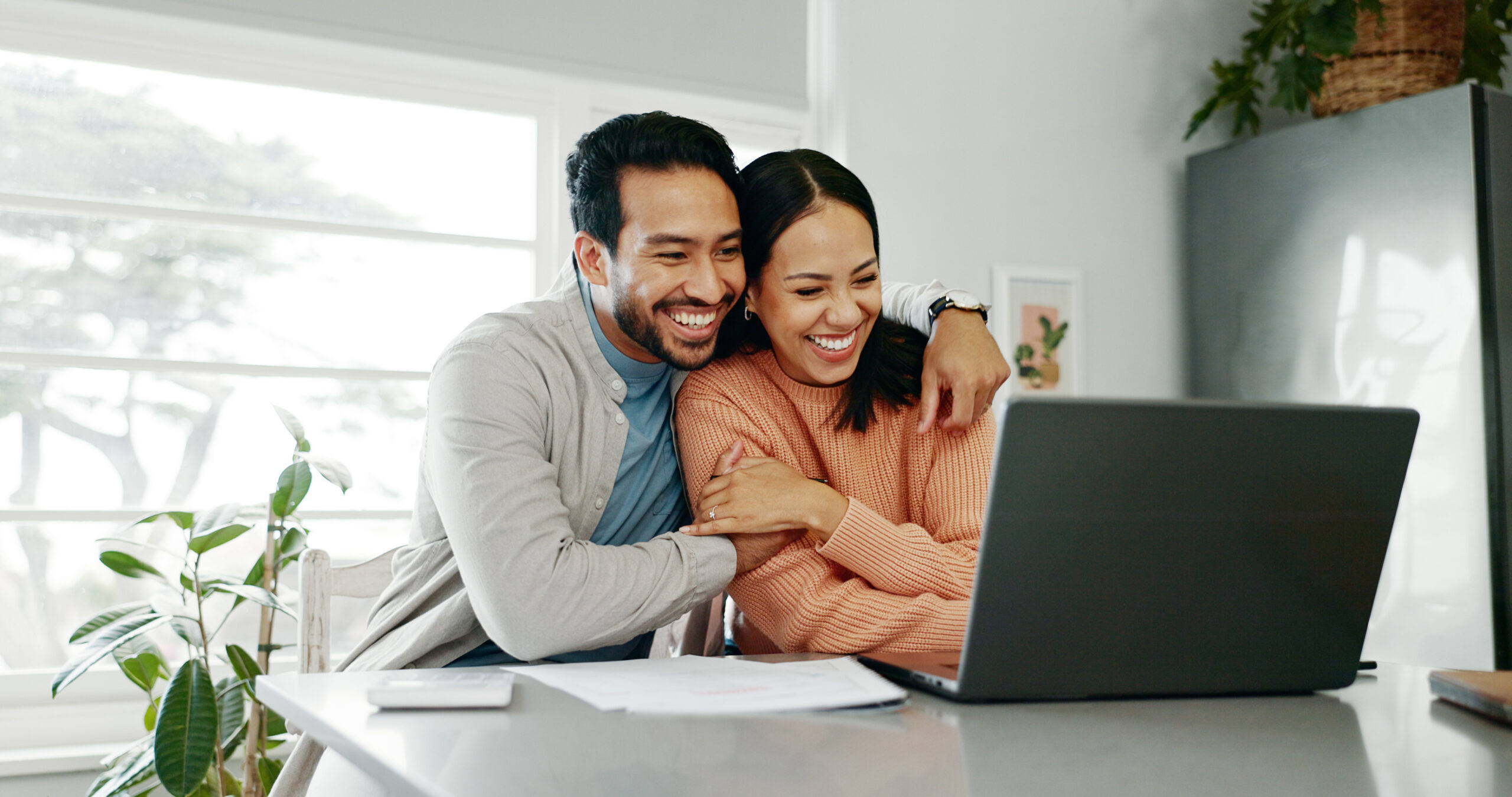 A man and a woman embrace while sitting at a desk, smiling, looking at their laptop.