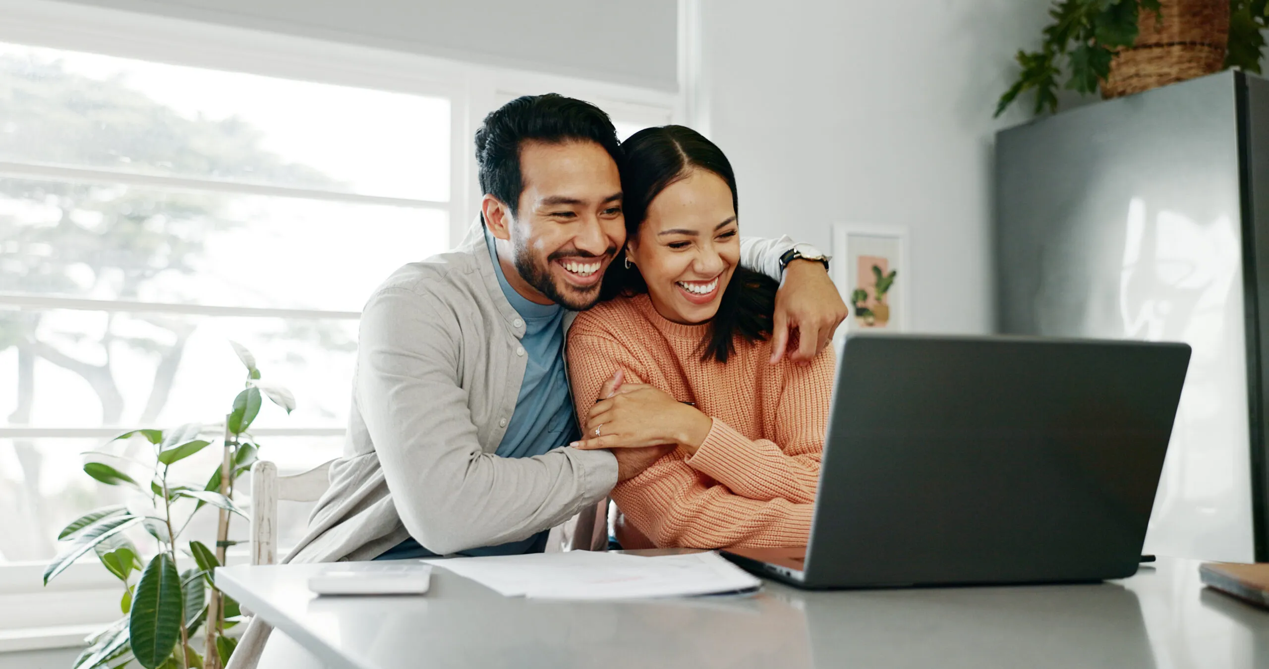 A man and a woman embrace while sitting at a desk, smiling, looking at their laptop.