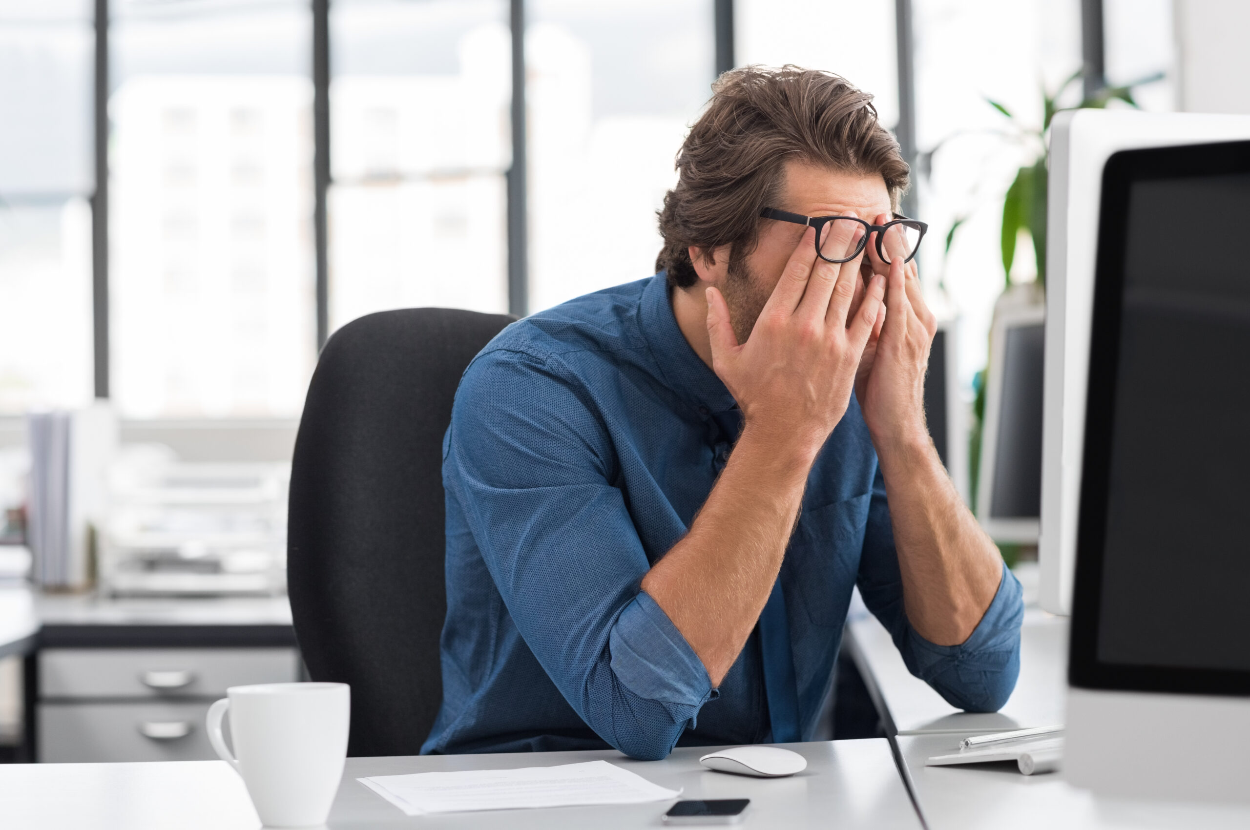 A man sits at his desk in a blue shirt and glasses while rubbing his eyes.