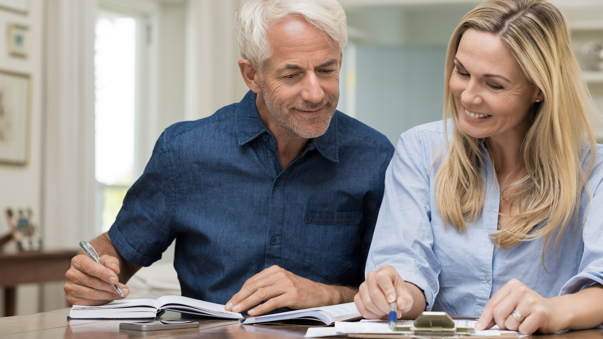 A man and woman sit at a desk, smiling while writing on a clipboard