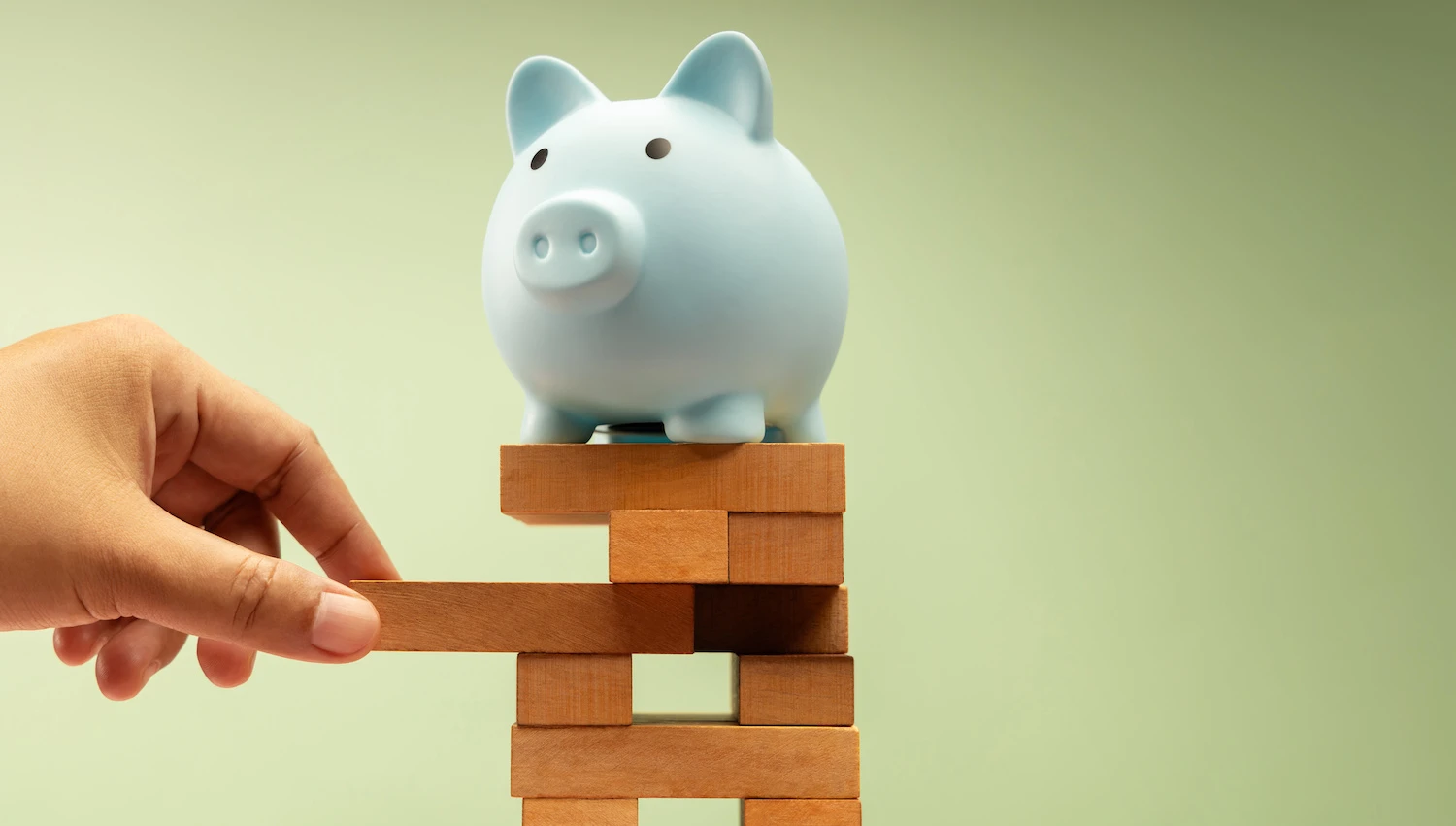 A light blue piggy bank sitting on top of a jenga tower while a hand pulls away one of the blocks.
