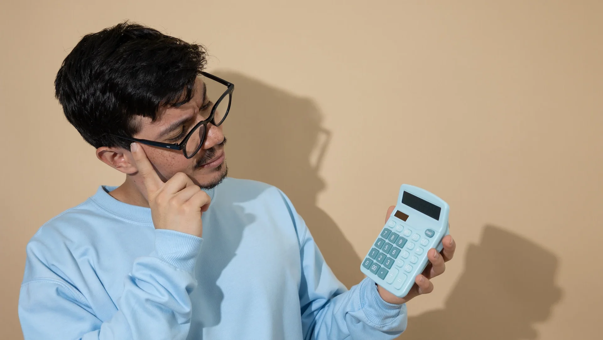 A man in a blue sweat shirt and glasses looks at a calculator he is holding in his left hand.