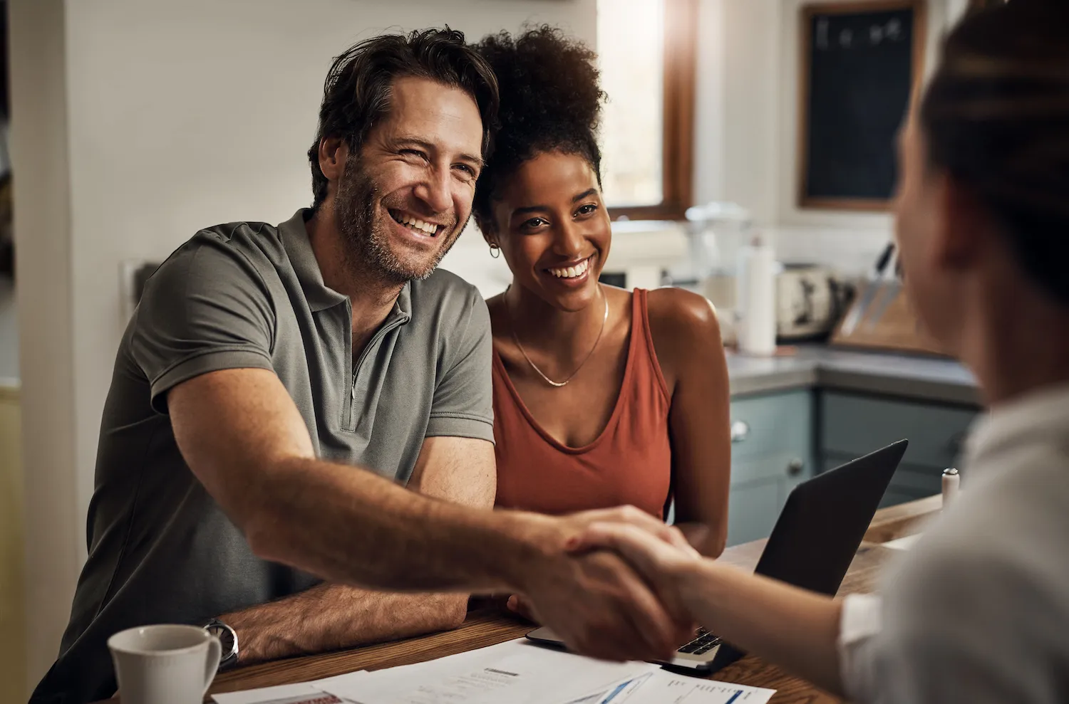 A man sits next to a woman at a desk while shaking hands with a man across the desk from him.