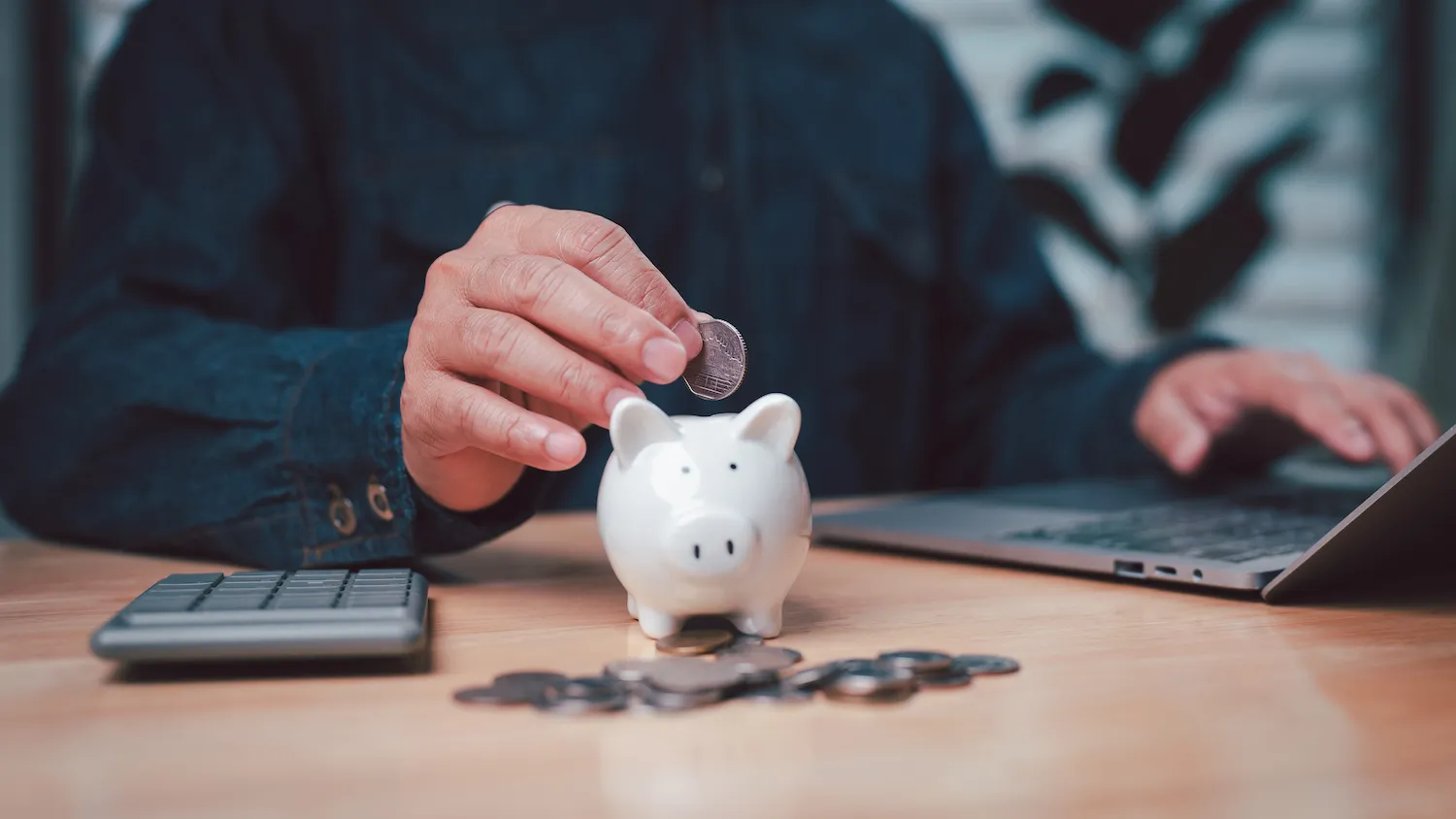 A man inserting a coin into a small white piggy bank sitting on a desk.