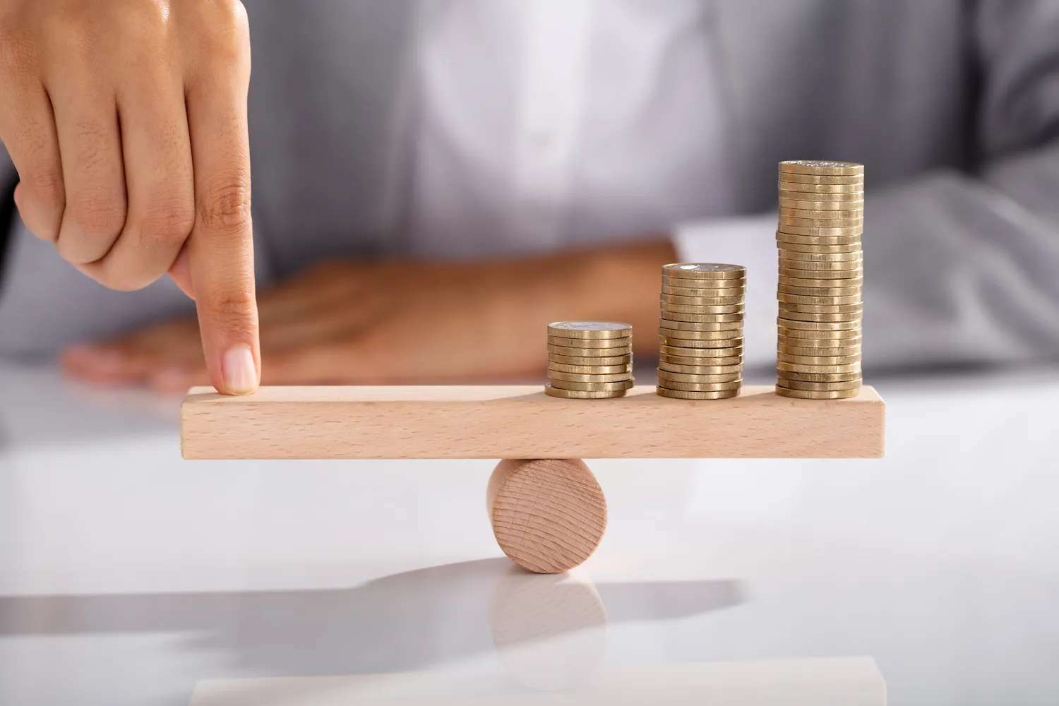 A person puts their finger on a scale with coins stacked on the other side.