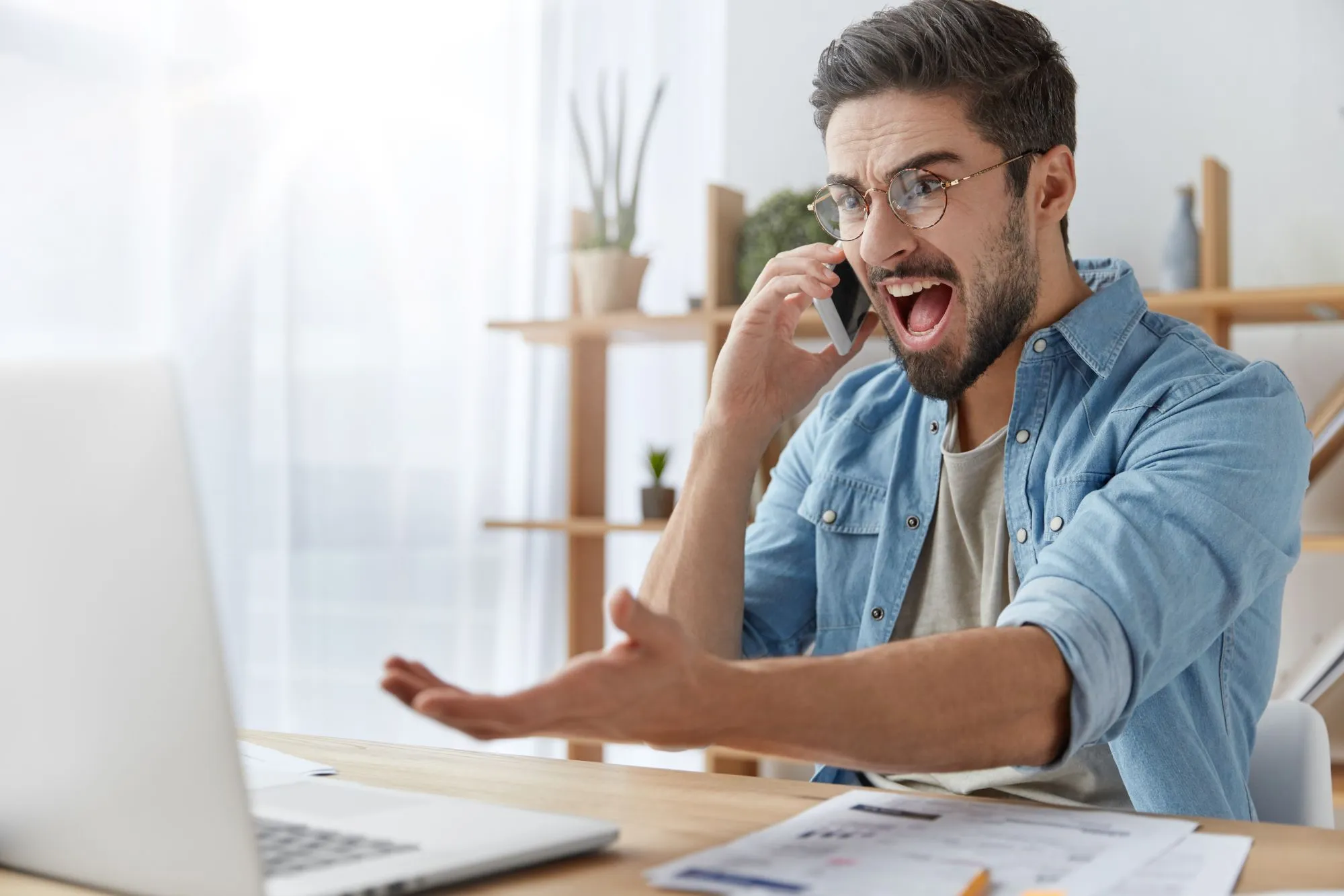 Man yells into cell phone while gesturing towards his computer while sitting at his desk.
