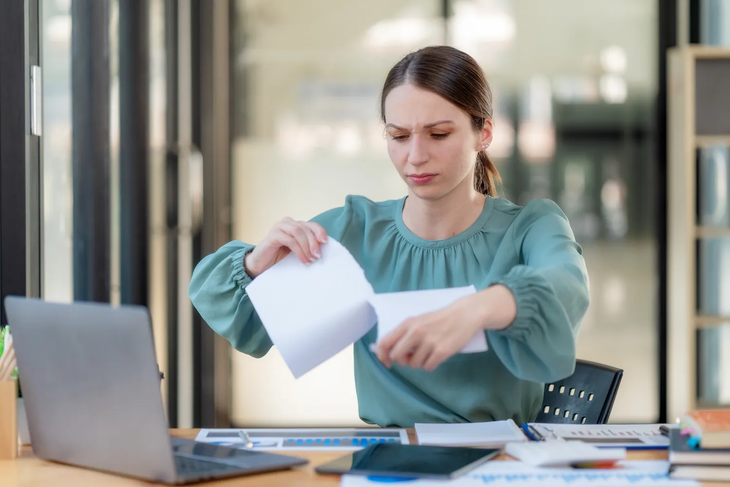 A woman in a green shirt rips up a piece of paper while sitting at her office desk.