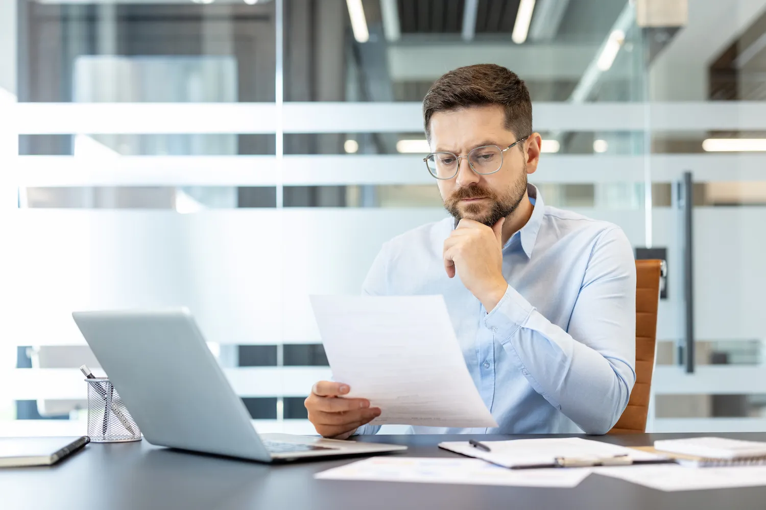 A man reads a paper while sitting at his desk in front of his computer.